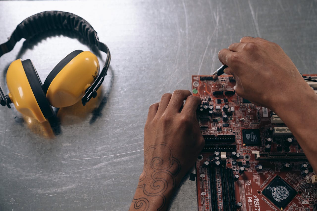 Home Hands repairing a motherboard on a metallic surface with yellow earmuffs beside it.